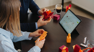 Two people working with colorful 3D models on a table with a tablet displaying a design app.