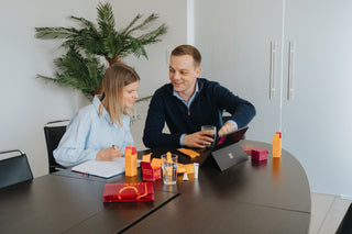 Two people sitting at a table with a laptop and various items, in an office setting.